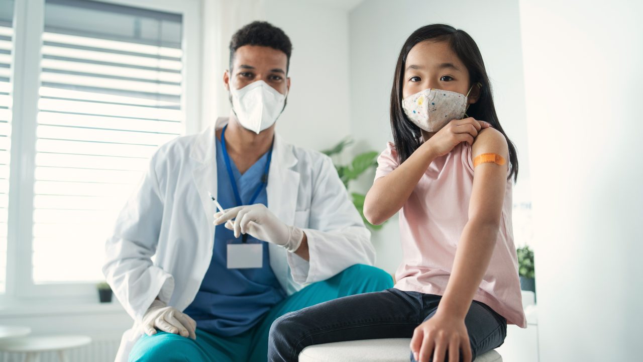 A nurse holds a needle next to a child with a bandage on their upper arm.