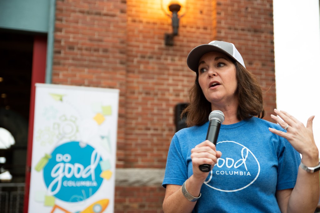 A person speaking into a microphone with a poster behind them reading "Do Good Columbia"