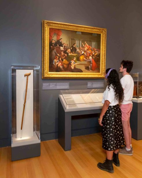 Two people wearing headphones and standing in front of a gallery wall with a painting and historical artifacts on display