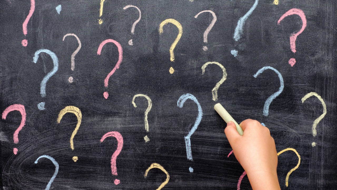 A hand holding up a piece of chalk to a chalkboard with question marks drawn on it