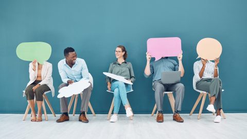 A group of people sitting in a lineup of chairs holding of sheets of paper shaped like speech bubbles