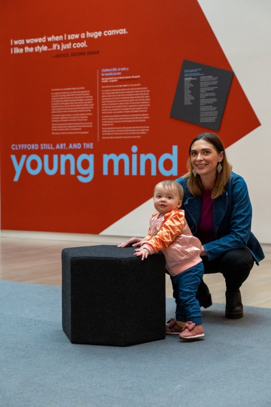 An adult and young child posing in front of the introductory wall text of the Clyfford Still, Art, and the Young Mind exhibition