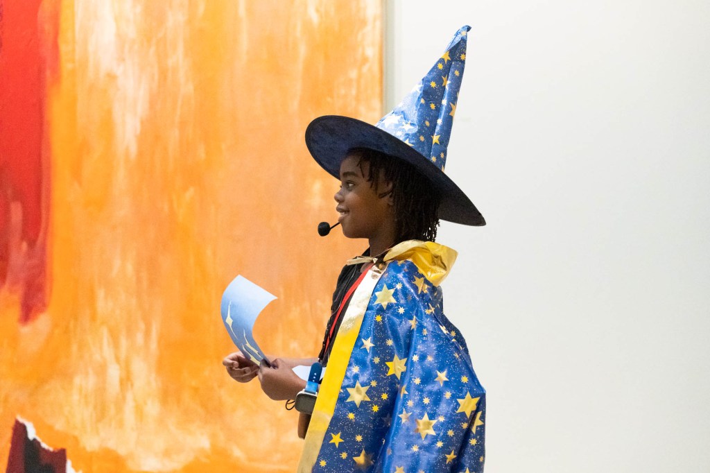 A young child wearing a wizard costume with a microphone and script standing in front of an abstract painting