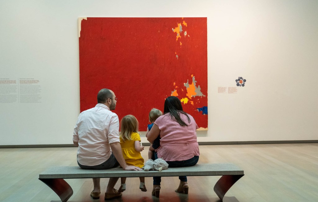 Two adults and two young children sitting on a bench in front of a large abstract painting