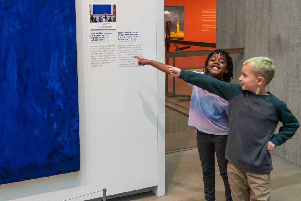 Two children smiling and pointing at wall text next to an abstract painting