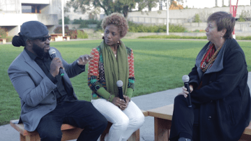 Two people sit on a bench opposite another person on a bench outside in a park. Each person holds a microphone. 