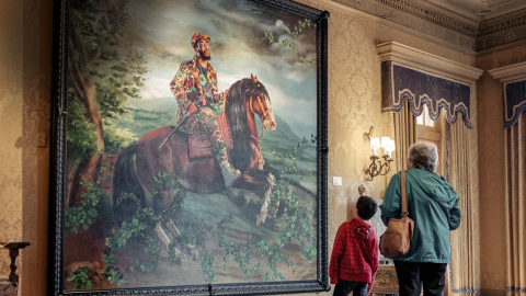 An adult and a child stand in front of a large, wall-sized, painting of a person on a horse that is rearing backward.