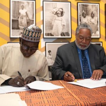 Two men sit behind a table with papers in front of them. They are both looking down and have pens in their hands to sign on the dotted line.