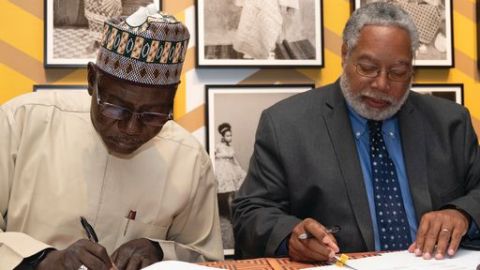 Two men sit behind a table with papers in front of them. They are both looking down and have pens in their hands to sign on the dotted line.