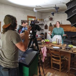 A person stands in front of a fireplace in historic costume speaking to a camera on the opposite side of the room.