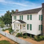 The exterior of a white colonial era house with a white picket fence surrounding it. 