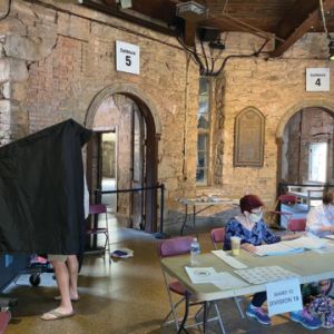 Several people sit at a folding table with another person behind a black curtained voting booth. 