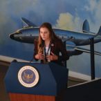 A person stands behind a podium with an airplane image in the background. 