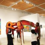 Several people view a sculpture in a museum gallery that looks like a melted metal sign painted orange.