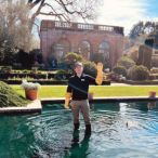 A person stands in a shallow pool of water waving at the camera outside in front of a historic structure in the background.