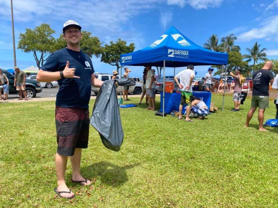 Dr. Hobbs standing on a grass field holding a trash bag and making the aloha hand sign, with other volunteers visible in the background