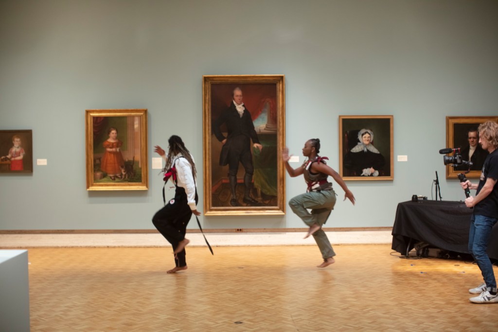 Two dancers doing a synchronized movement in a gallery of portrait paintings with a videographer trailing them