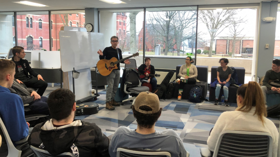 A room of people seated in a circle around someone holding a guitar