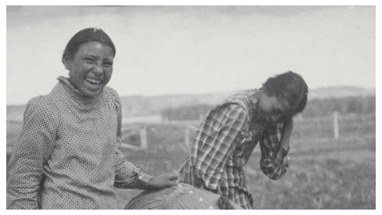 Three young women standing in a field (one is bent over forward) laughing for the camera. All are wearing long shirt-waist dresses.