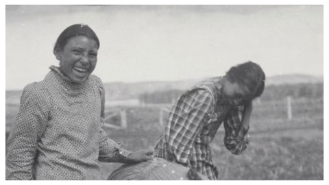 Three young women standing in a field (one is bent over forward) laughing for the camera. All are wearing long shirt-waist dresses.