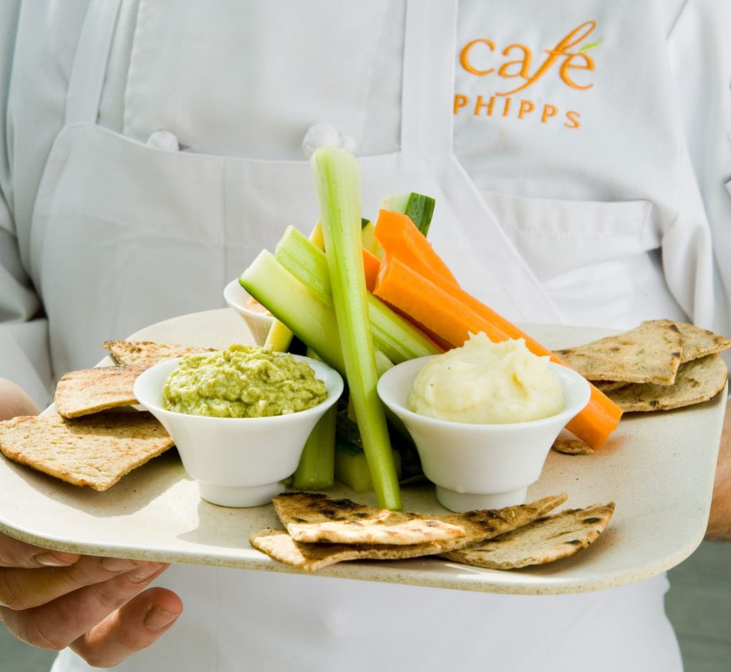 A chef wearing a uniform reading "Cafe Phipps" and holding a plate of flatbreads, vegetables, and spreads
