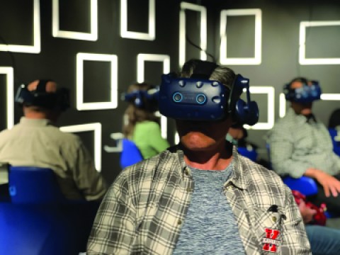 A person stands wearing a VR headset in front of other people in the background of an exhibition.