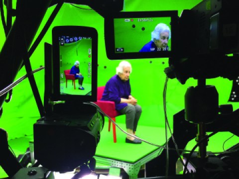 A woman sits in a red chair wearing a blue suit in a green screen theater setting. Several pieces of recording equipment sit in the foreground recording the woman in the chair. 