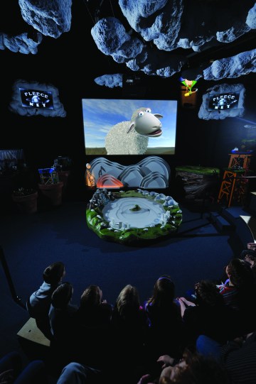 An audience watches a talking cartoon sheep on a screen in front of them. 