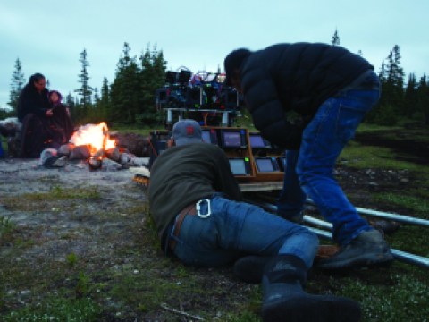 Two people crouch next to film equipment in front of a bonfire in the great outdoors. 