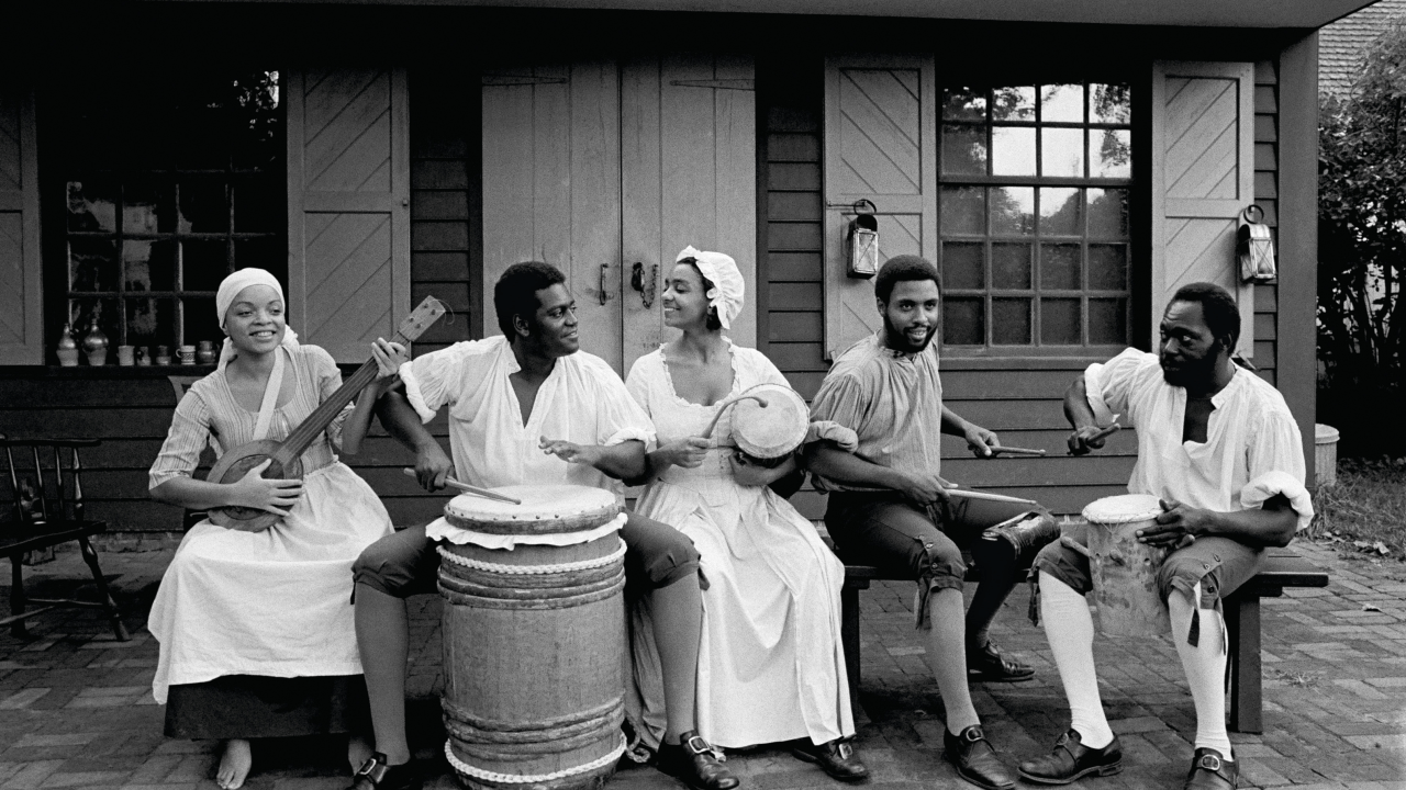 A group of actor dressed in colonial garb sit in front of a cabin playing various instruments.