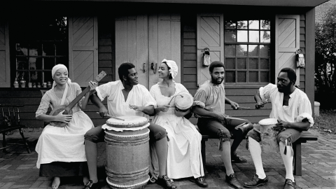 A group of actor dressed in colonial garb sit in front of a cabin playing various instruments.