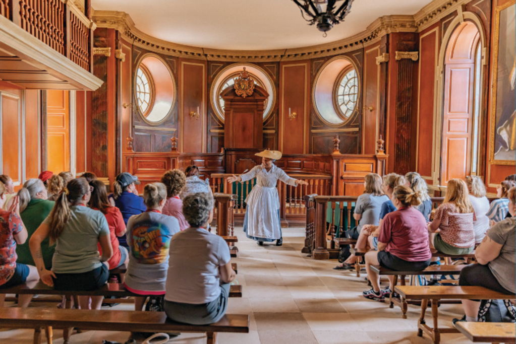 An audience of people sit in a historic room watching a performer wearing a colonial dress and hat give a speech.