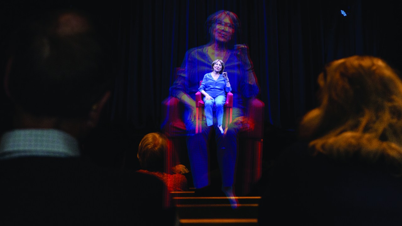 A seated woman in a blue suit is holographically projected forward on a stage.