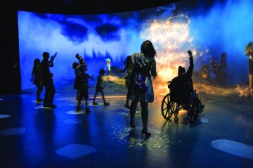 Several people, including one person in a wheelchair, dance around a bonfire.