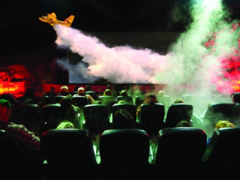 A audience in a theater watches as an airplane releases smoke suppressant over a fire.
