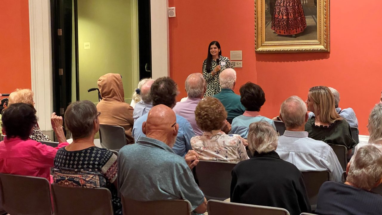 An educator speaking in front of a seated audience in front of a painting