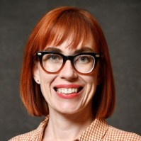 A white woman with straight red hair and wearing dark rimmed glasses and a red and white checkered button down shirt.