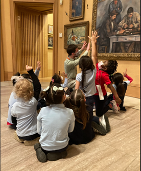 A gallery educator speaking to a group of young students seated on the floor in front of a painting.