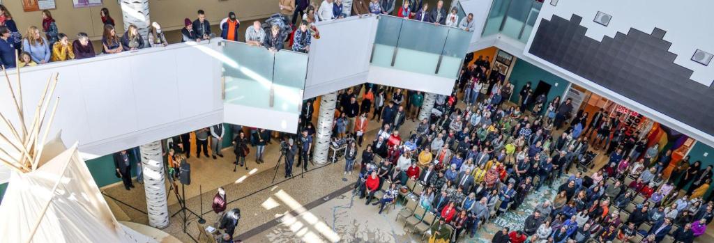 An aerial view of a public event in a museum space where a person in traditional Indigenous clothing stands on stage