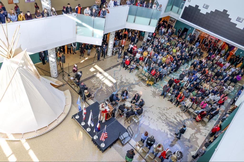 An aerial view of a public event in a museum space where a person in traditional Indigenous clothing stands on stage