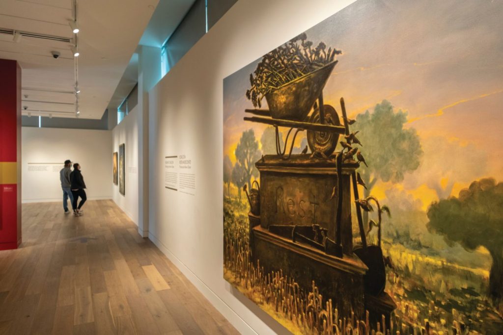 An exhibit panel showing a painting of a statue of a wheelbarrow on top of a plinth. 