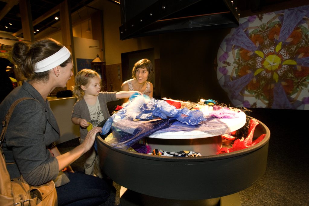 Three people stand at a circular table with various pieces of colorful fabric that they can interchange to make a kaleidoscope. 