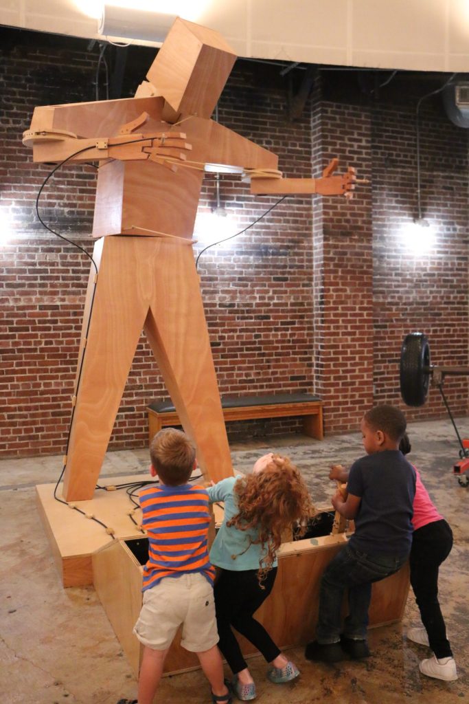 Several children stand at an exhibition feature with a large oversized wooden figure above them. 
