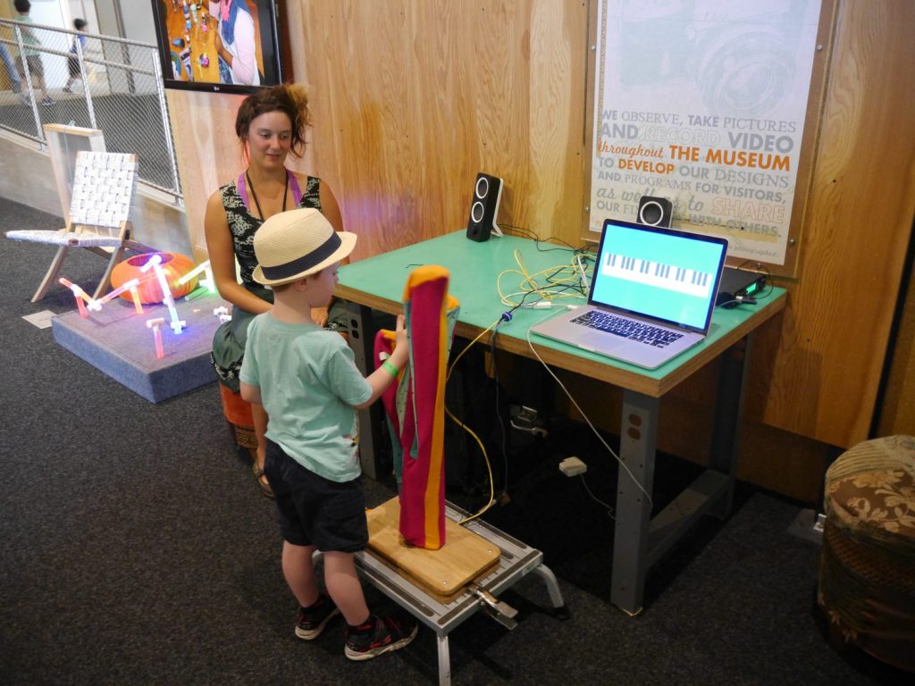 A child and an adult stand at an interactive station with an open laptop with a piano app playing. 