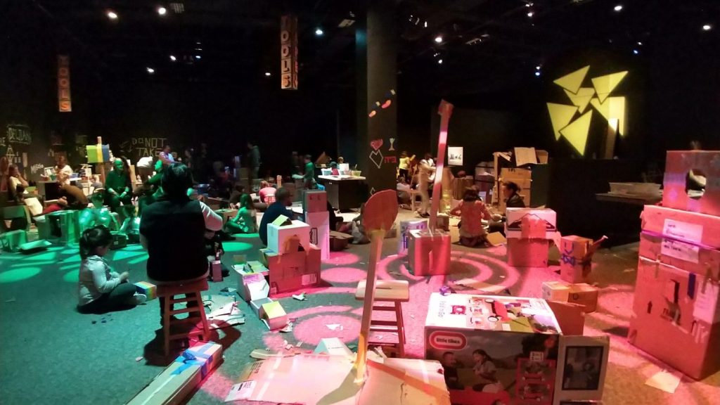 Several children sit, lean, play in a forest of cardboard boxes.