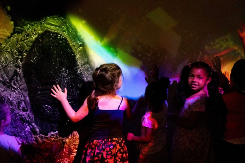 A couple of children stand with their backs to the camera touching various elements of an interactive exhibit feature.