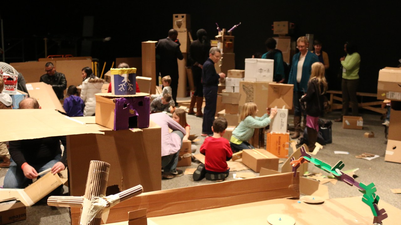 Several children sit, lean, play in a forest of cardboard boxes.