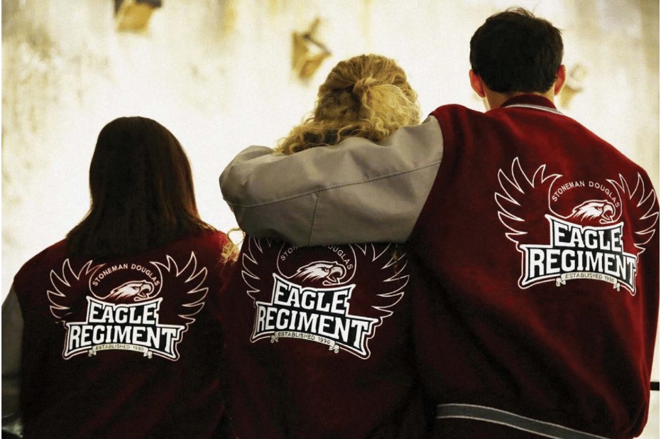 Three people stand with their backs to the camera with red letterman jackets on that say Eagle Regiment with a spread eagle embroidered on them.