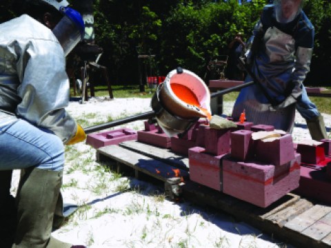 Two men stand next to a large vat of red or pink liquid being poured into a mold. 