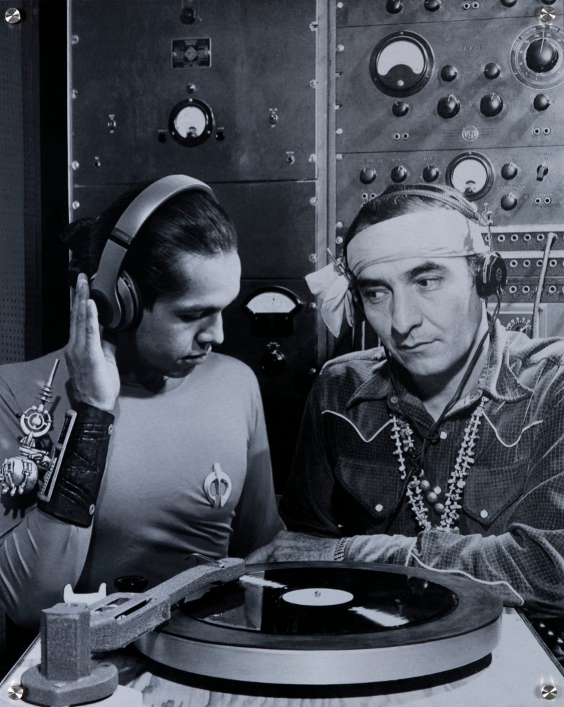 A black and white photograph of two Indigenous people, one wearing a sci-fi costume and the other Western clothing, listening to a vinyl record next to a control panel.